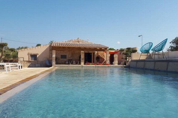A swimming pool with a stone building in the background, a white fence, and two blue chairs.
