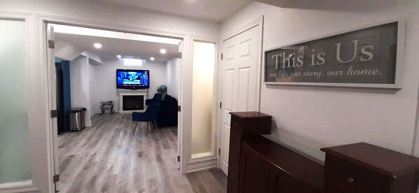 A hallway with a television, a blue chair, and a sign that says "This is Us". - Adorable 2 bedrooms, 2 washrooms, 2 parking spots apartment (Newmarket)