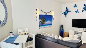 A living room with a white dining table, a grey sofa, and a TV on a cabinet. There is a painting of a beach scene on the wall. - Your Own Private Rooftop Paradise — Hammocks, Ocean Views & Steps from the Beach (Playa Los Cerritos)