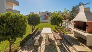 An outdoor patio with a table and benches, surrounded by greenery and a barbecue area. - Apartments Anamarija - One Bedroom Apartment with Balcony (Privlaka)