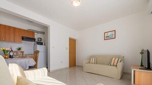 A living room with a sofa, a dining table with chairs, a kitchen area, and a television on a stand. - Apartments Anamarija - One Bedroom Apartment with Balcony (Privlaka)