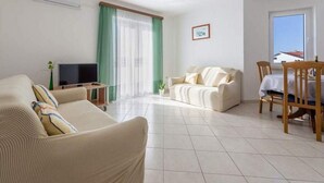 A living room with a sofa, a TV, a dining table with chairs, and a window with curtains. - Apartments Anamarija - One Bedroom Apartment with Balcony - 2 (Privlaka)