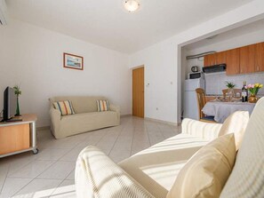 Uma sala de estar ampla com um sofá, uma mesinha com uma planta e uma cozinha visível por uma porta aberta.