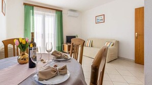 Zona pranzo con tavolo apparecchiato per due, una TV sullo sfondo e un divano nella stanza adiacente.