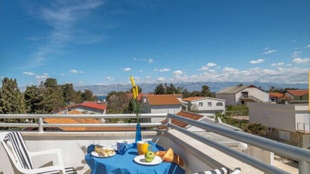Un balcone con un tavolo apparecchiato per due, vista su una zona residenziale con case e montagne sullo sfondo.