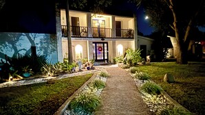 A two-story building with a central entrance, surrounded by a well-maintained garden with various plants and a gravel pathway leading to the door. - Tropical Garden Oasis  • Birdwatcher's Paradise • Spacious Harlingen Retreat (Harlingen)