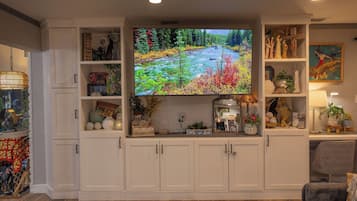 Een woonkamer met een grote flatscreen televisie, bevestigd aan een wit meubel, omringd door planken met decoratieve objecten en planten.