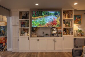 A living room with a large flat-screen TV mounted on a white cabinet, surrounded by shelves with decorative items and plants. - Tropical Garden Oasis  • Birdwatcher's Paradise • Spacious Harlingen Retreat (Harlingen)
