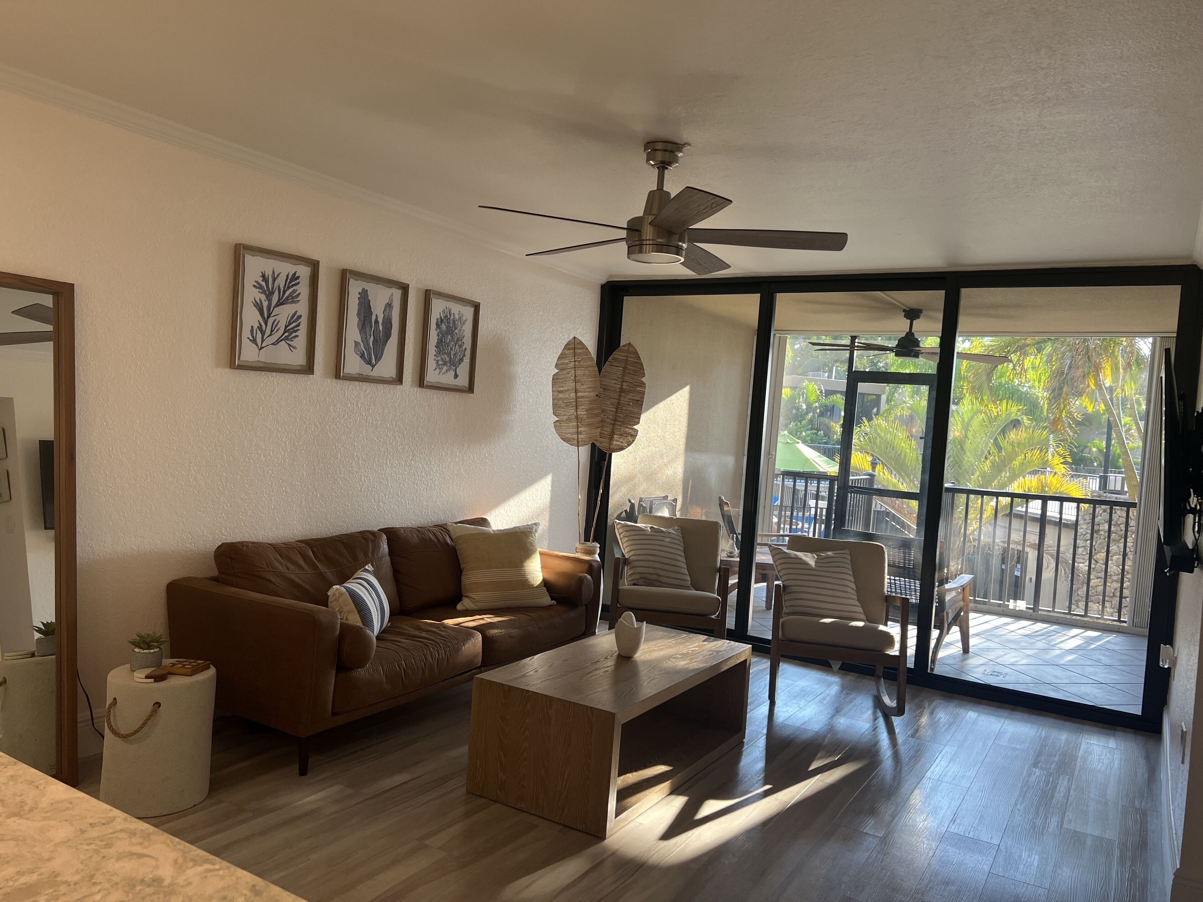 A living room with a brown leather sofa, a wooden coffee table, and a large window with a view of a balcony and greenery.