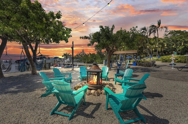 A beachside seating area with turquoise Adirondack chairs arranged around a central fire pit, overlooking a marina and a sunset.