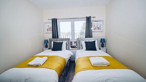 Ein Doppelzimmer mit zwei Betten, jeweils mit weißer und gelber Tagesdecke und passenden Kissen. Zwei Nachttische mit Lampen sind vorhanden, sowie ein Fenster mit Vorhängen.