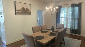 Ein Esszimmer mit einem Holztisch, sechs Stühlen, einer Kristallleuchter, einem gerahmten Bild an der Wand und einem Fenster mit Vorhängen.
