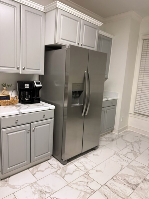 A kitchen with grey cabinets, a stainless steel refrigerator, a coffee maker, and marble flooring.