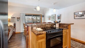 Een keuken met een granieten aanrechtblad, houten kastjes en een zwarte oven. Er zijn twee hanglampen, een raam met uitzicht op bomen en een deur die leidt naar een andere kamer.