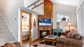 Een gezellige woonkamer met een open haard, een aan de muur bevestigde televisie en een trap naar een andere kamer.