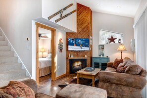 Ein gemütliches Wohnzimmer mit Kamin, einem an der Wand befestigten Fernseher und einer Treppe, die in ein weiteres Zimmer führt.
