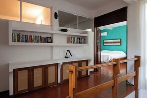 A room with a wooden staircase, a white wall with shelves containing books, a bed with a white cover, and a teal-colored wall.