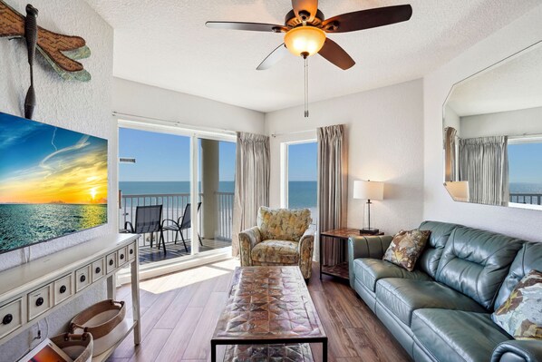 A spacious living room with a large window overlooking the ocean, a ceiling fan, a leather sofa, and a coffee table.