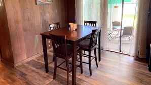 A dining area with a wooden table and chairs, a hanging light fixture, and a view through a glass door to an outdoor patio.