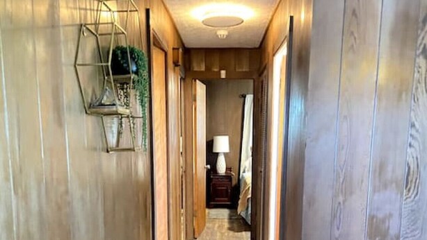 A hallway with wooden walls and a wooden floor, leading to a room with a lamp and a framed picture on the wall.