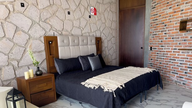 Un dormitorio con pared de piedra, pared de ladrillo, cama con ropa de cama oscura, mesita de noche con velas y una pequeña mesa con un florero.
