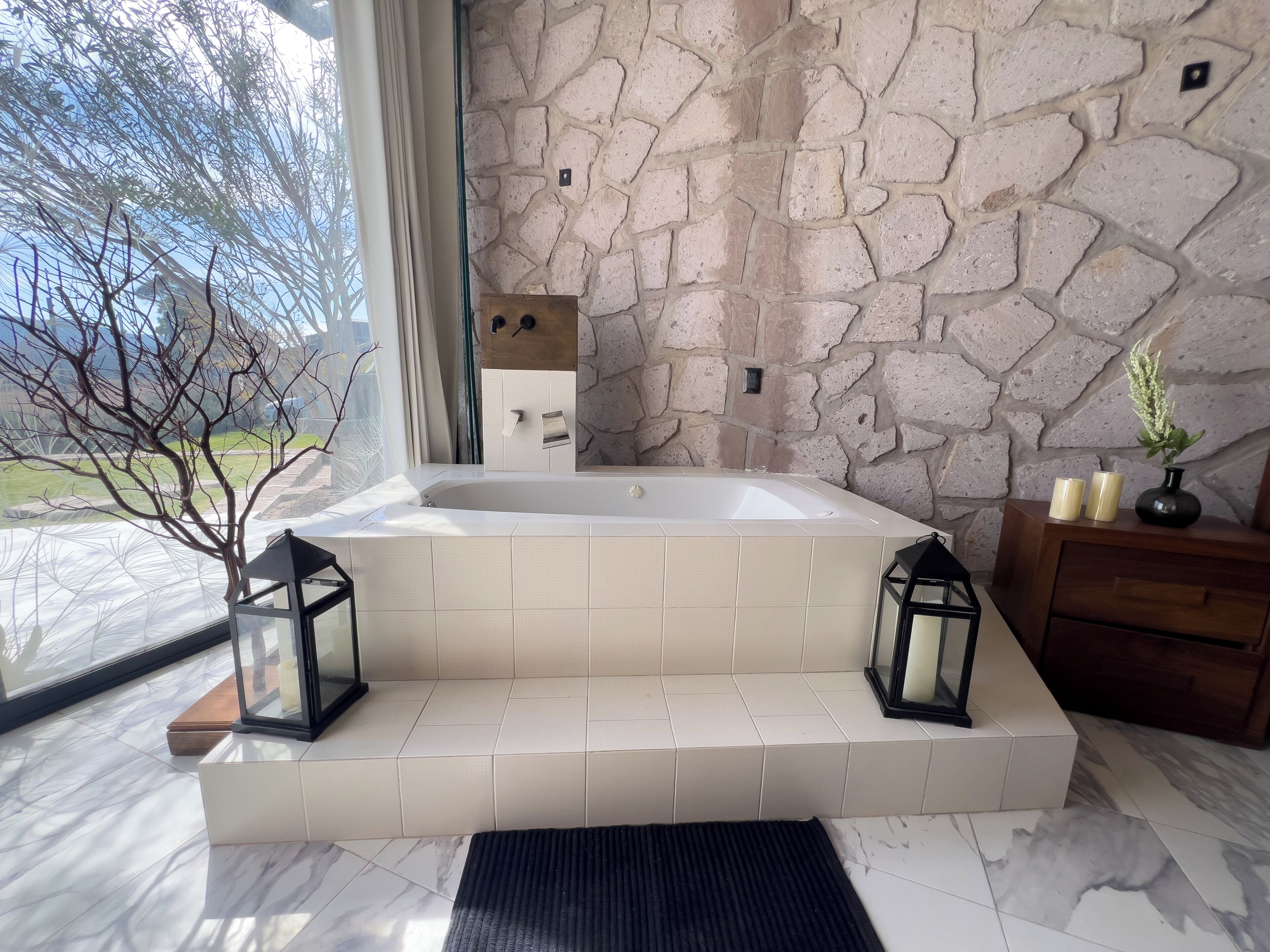 Un baño moderno con pared de piedra, una bañera grande y un tocador de madera con velas.