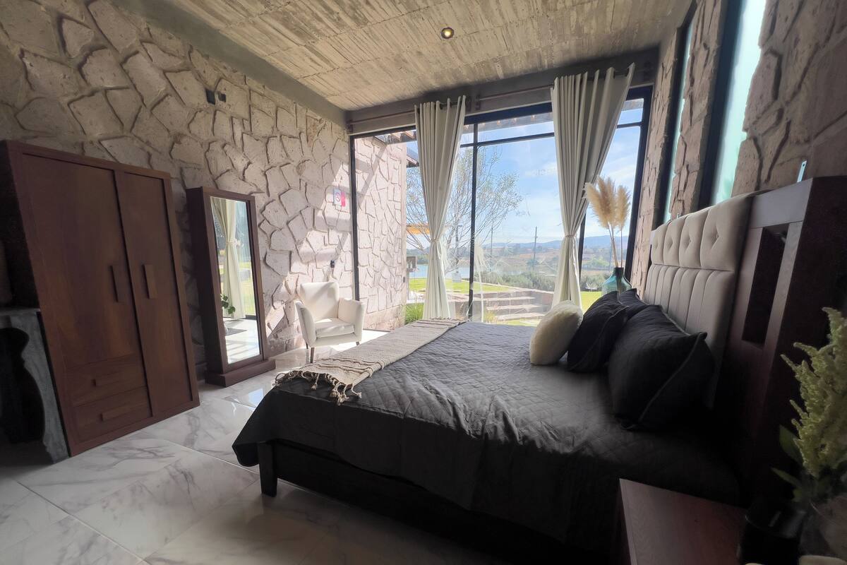 Un dormitorio con pared de piedra, cama con ropa oscura, armario de madera y vista al exterior.