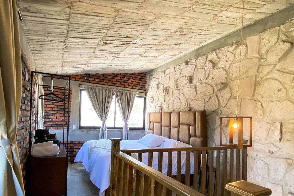 Una habitación con techo de madera, pared de piedra, cama con ropa blanca, lámpara colgante y baranda de madera.