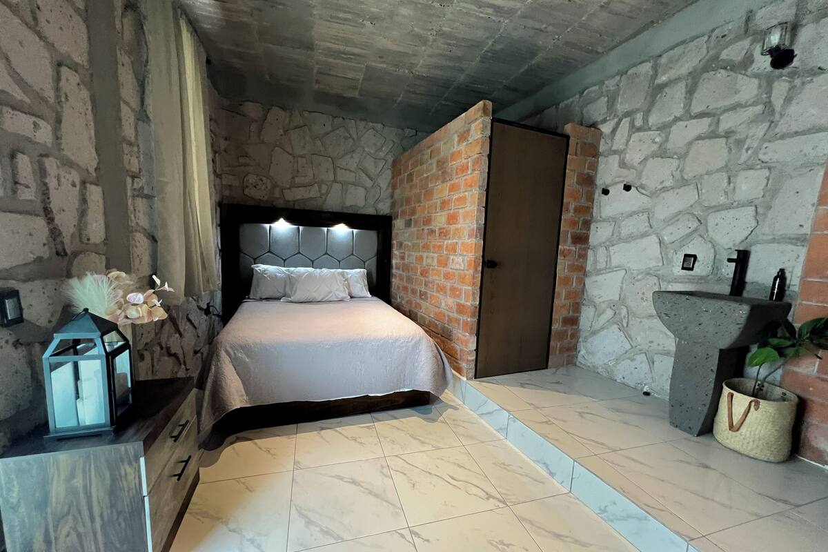 Un dormitorio con pared de piedra, una cama con ropa de cama blanca, una mesita de noche, una lámpara y una pequeña planta.