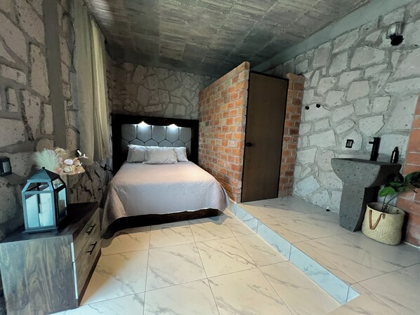 Un dormitorio con pared de piedra, una cama con ropa de cama blanca, una mesita de noche, una lámpara y una pequeña planta.