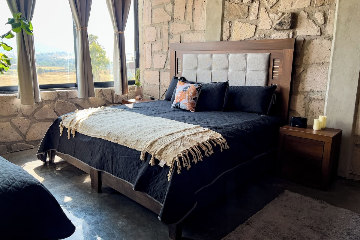 Un dormitorio con pared de piedra, cama con colcha azul oscuro, cabecero de madera y mesita de noche con velas.