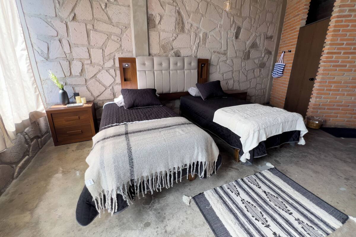 Una habitación con dos camas, pared de piedra, cabecero de madera, mesita de noche, una pequeña mesa con un florero y una alfombra estampada.