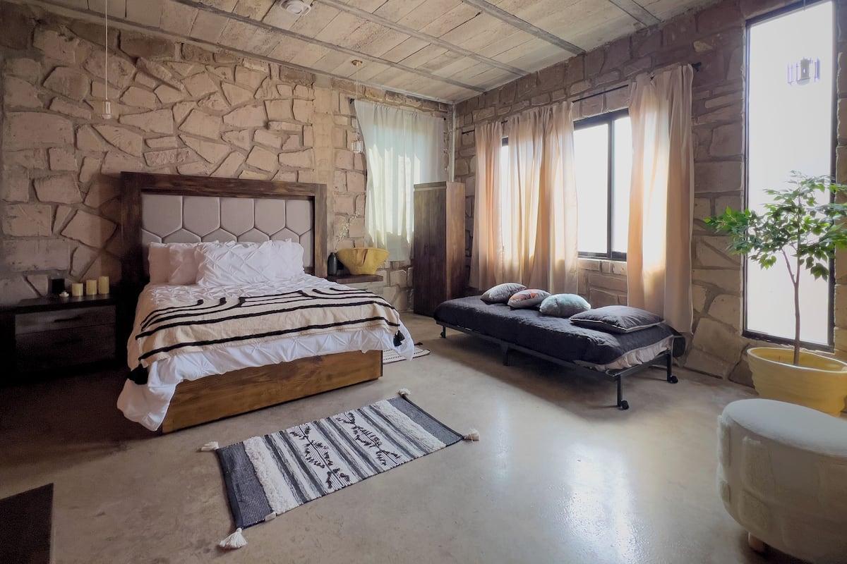Un dormitorio con pared de piedra, cama de madera, sofá cama, mesita de noche, lámpara, ventana con cortinas y una planta en maceta.