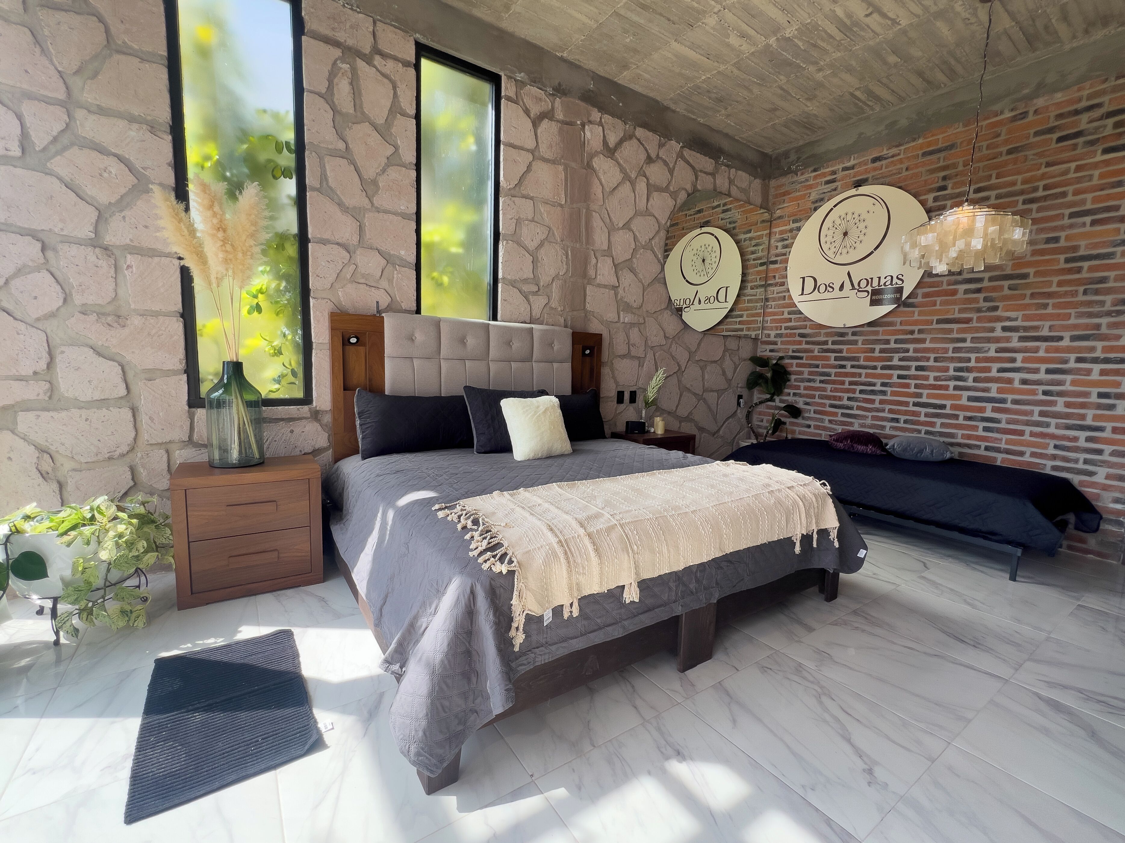 A bedroom with a stone wall, a bed with a grey bedspread, a nightstand, a bedside table, and a hanging light fixture.