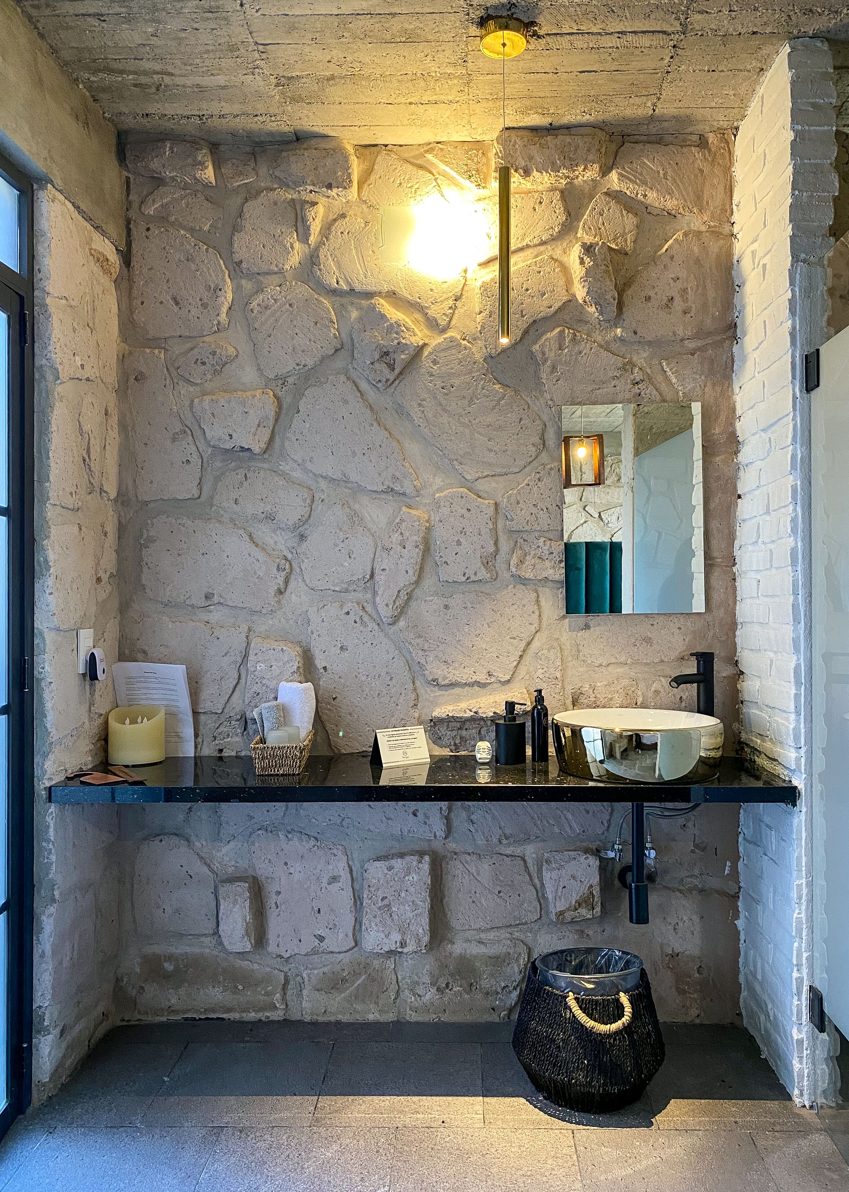 Un baño con pared de piedra, encimera negra, espejo, lavabo, perchero y cubo de basura.