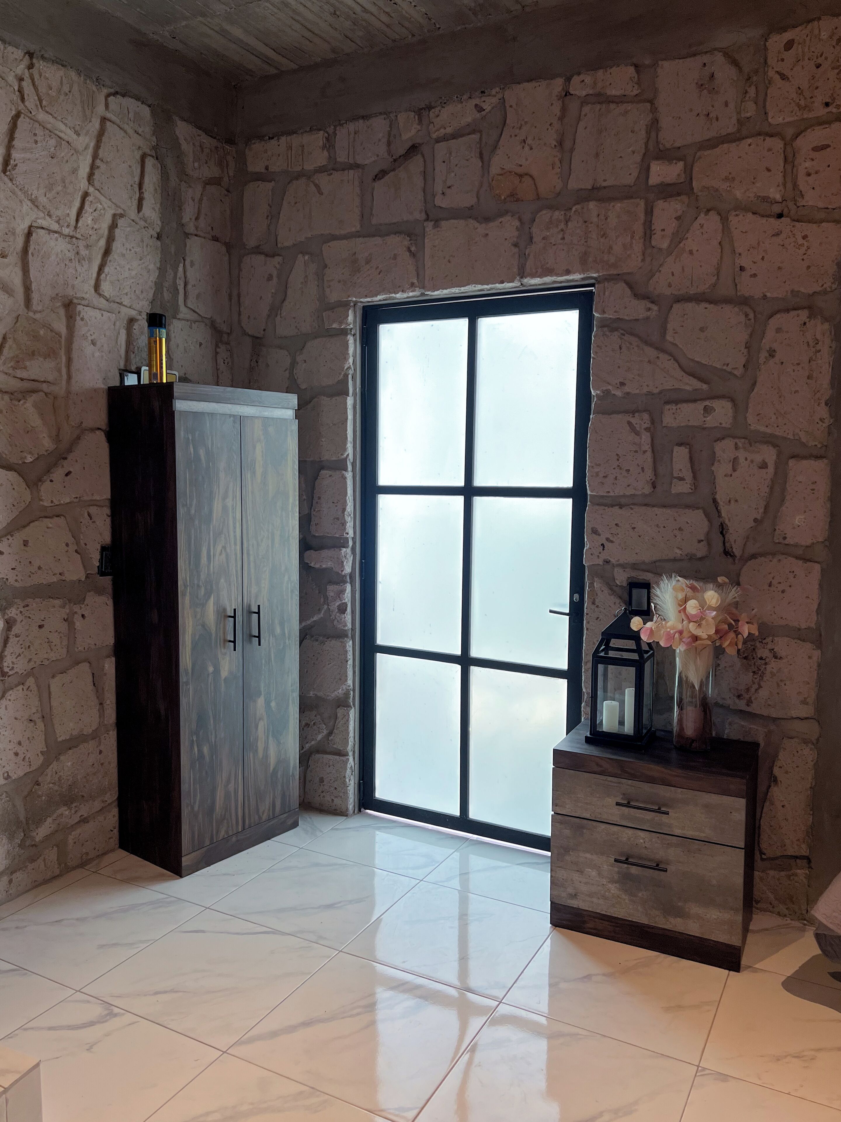 Una habitación con pared de piedra, un armario de madera, una puerta de cristal y un pequeño mueble de madera con un jarrón de flores.