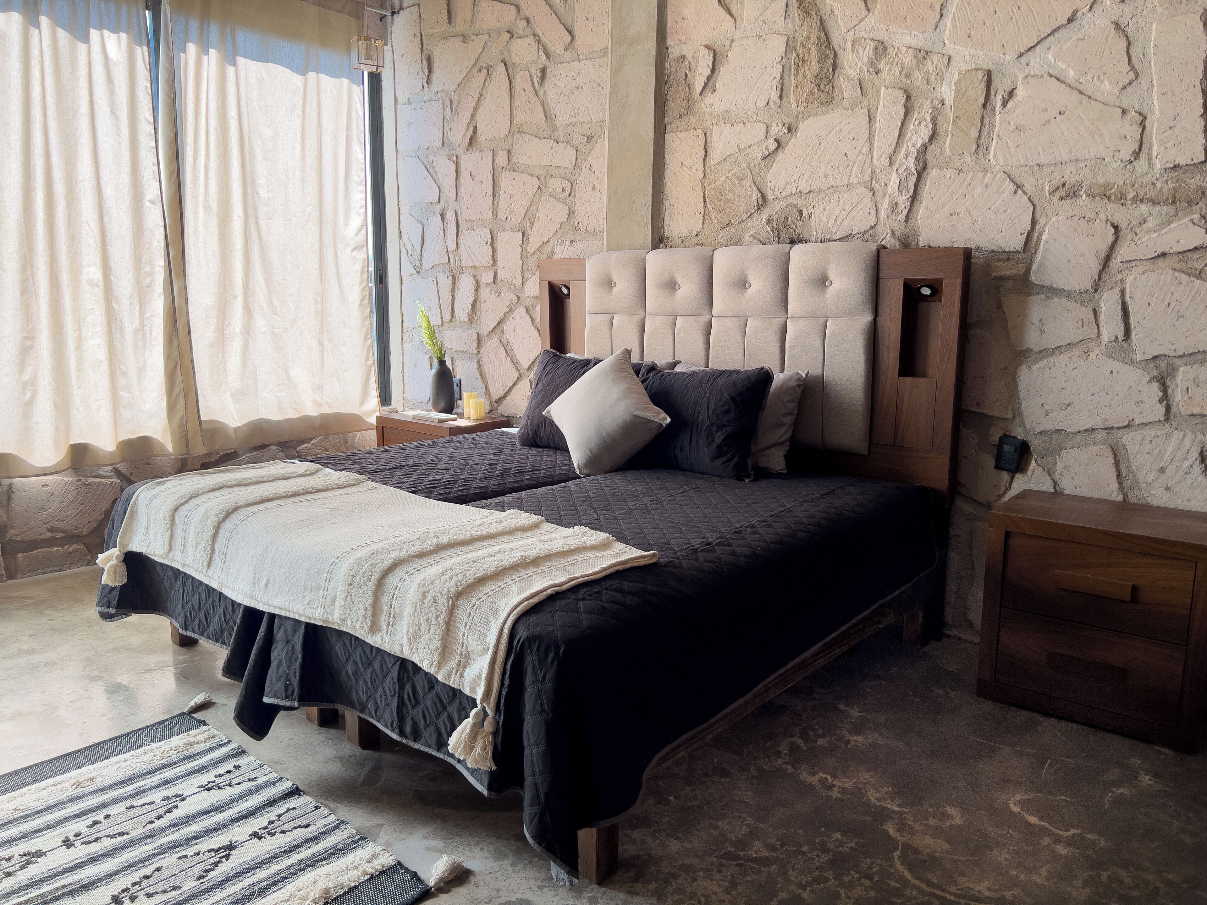 A bedroom with a stone wall, a bed with dark bedding, a bedside table with a vase and candles, and a window with curtains.