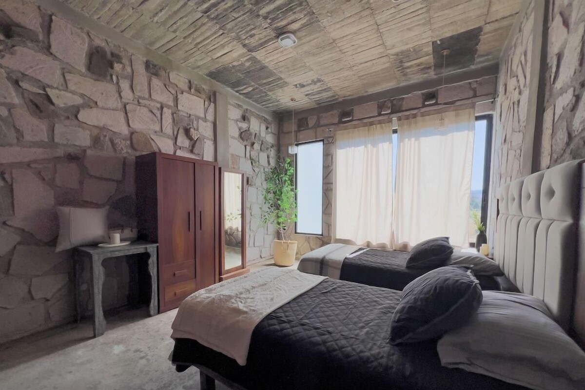 Un dormitorio con paredes de piedra, una cama, una mesita de noche, un armario y una ventana con cortinas.