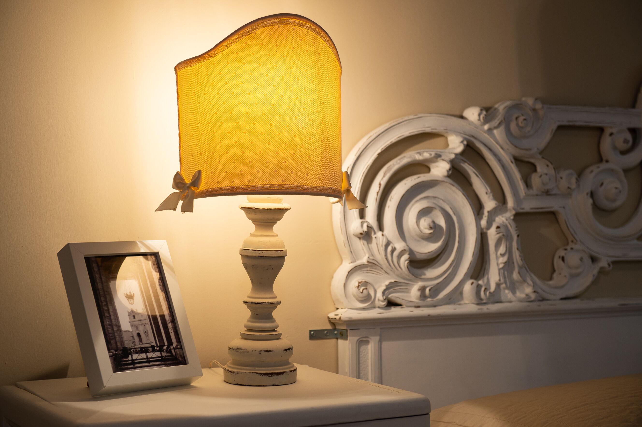 Une table de chevet avec une photo encadrée, une lampe blanche avec un abat-jour jaune et une tête de lit blanche décorée.
