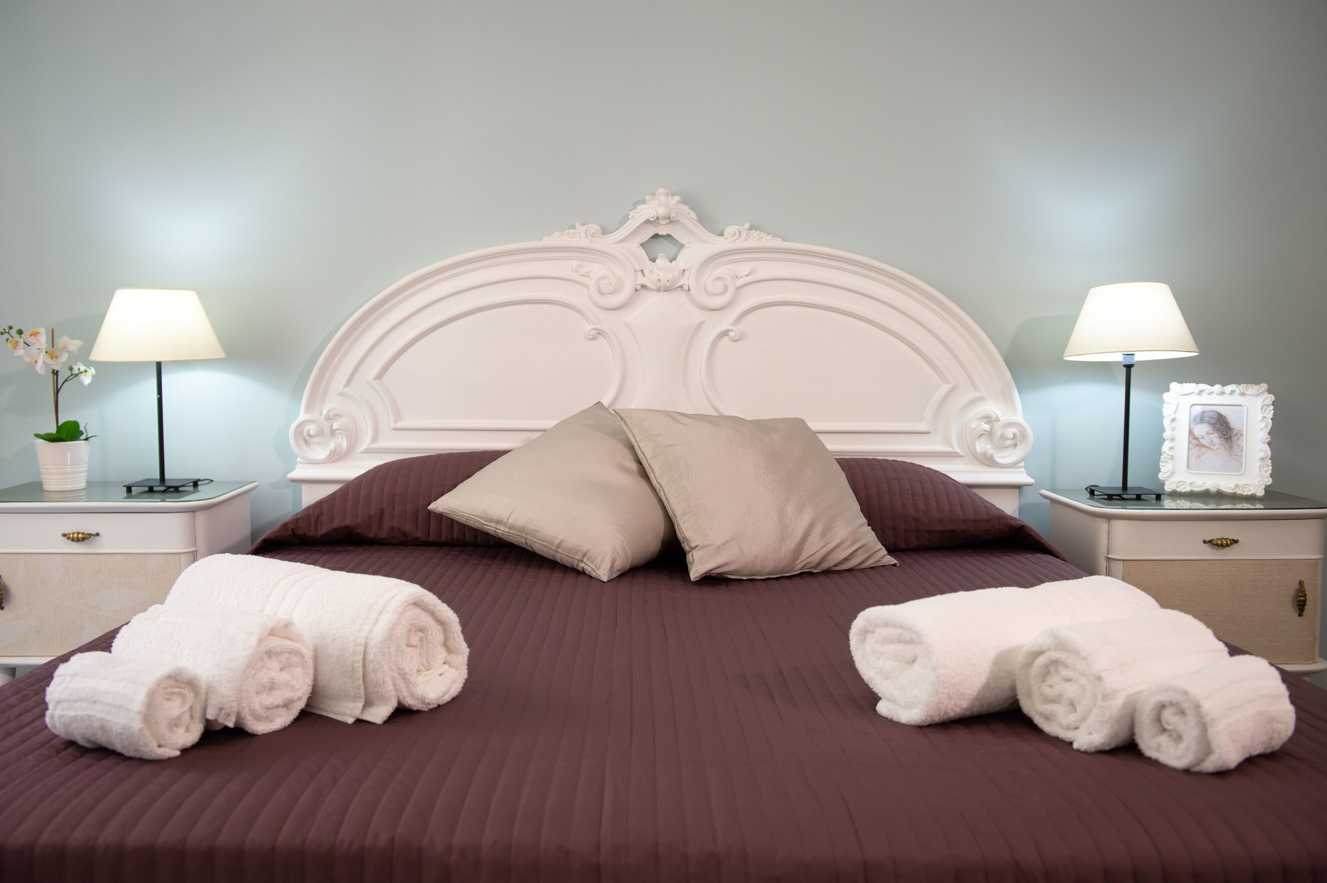 A neatly made bed with brown bedding and two beige pillows, flanked by two nightstands with lamps and a framed picture.