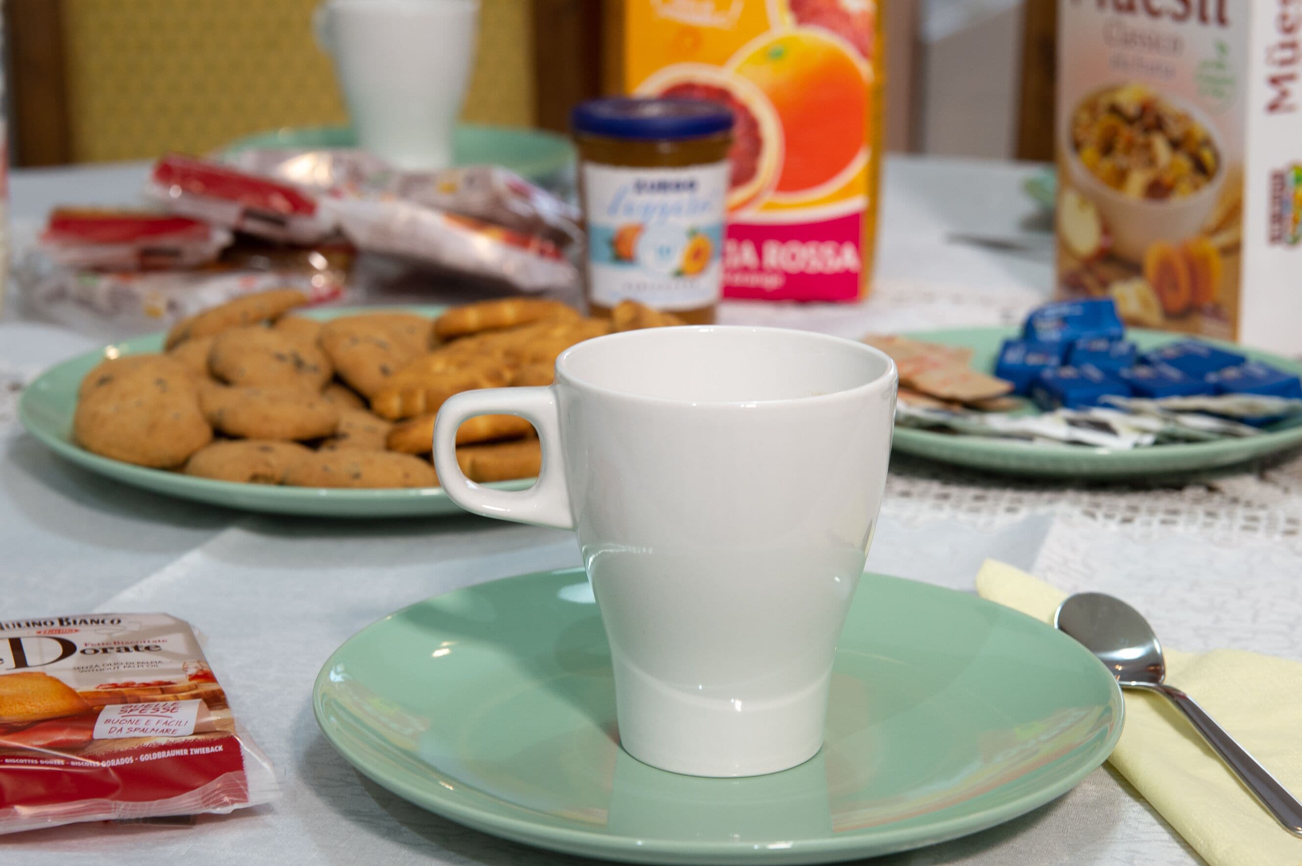 A table with a white mug on a green saucer, a plate of cookies, and various food items including a jar of marmalade and a box of cereal.