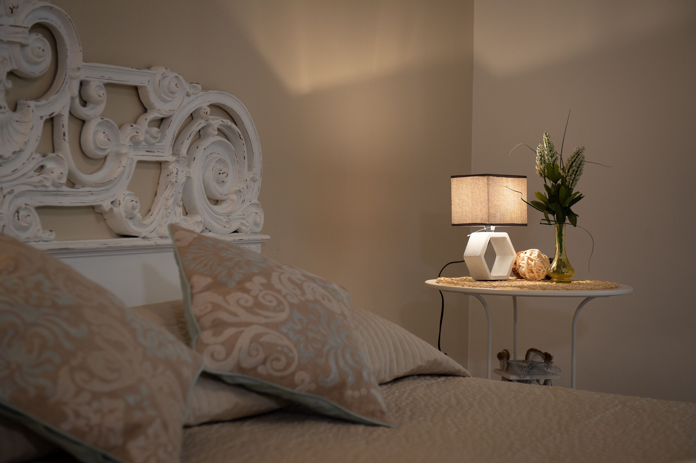 A neatly made bed with patterned pillows, a decorative headboard, a small table with a lamp and a vase with plants, and a wall-mounted shelf with a decorative item.