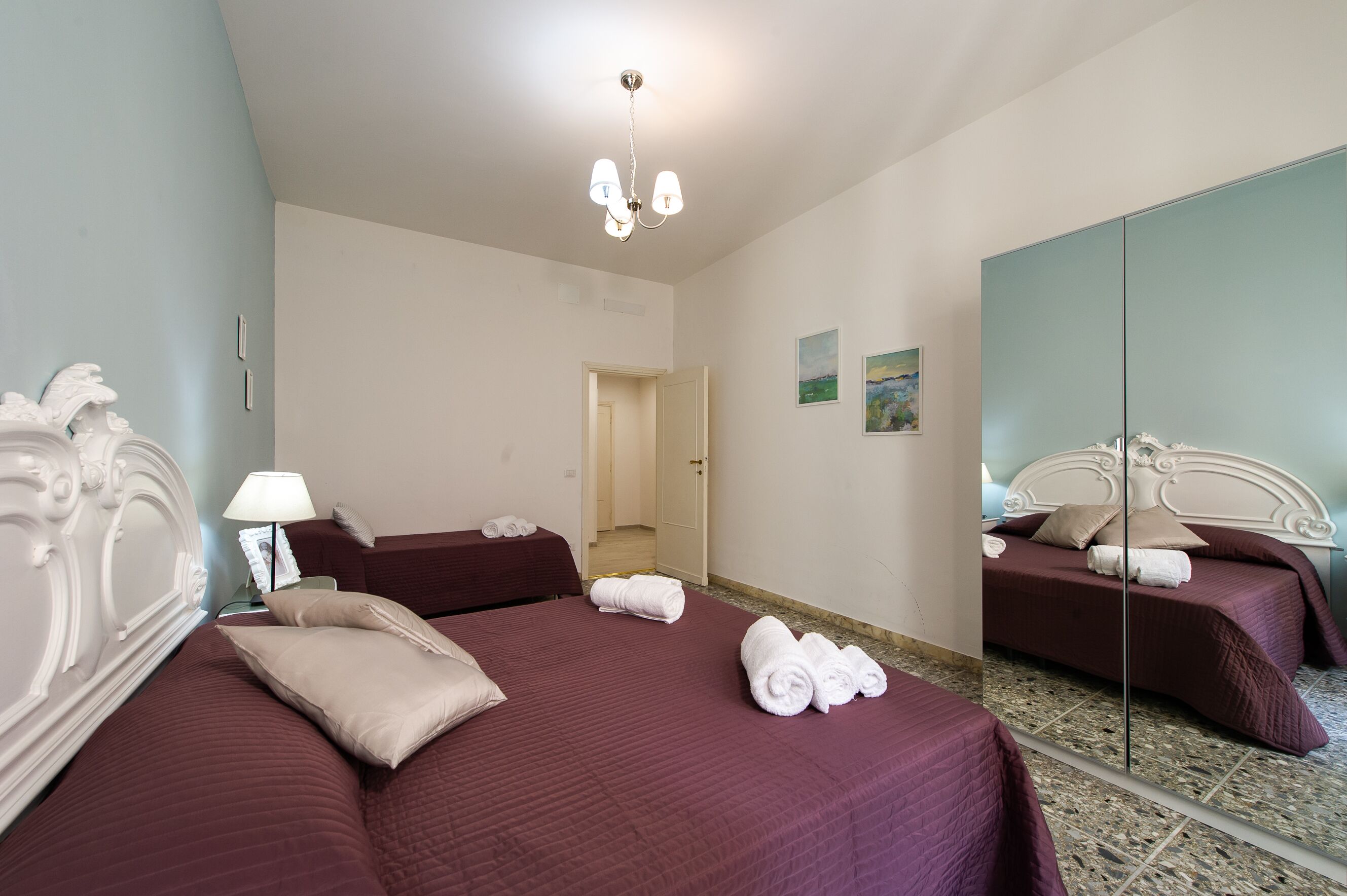A hotel room with two beds, a chandelier, a mirror, and a door leading to another room.