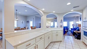 A spacious kitchen with white cabinetry and a large island, connected to a dining area with blue chairs and a TV.