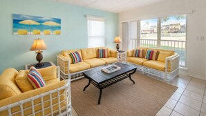 A living room with a yellow sofa set, a coffee table, and a painting of umbrellas on the wall. - Beach View Condo 106 By The Beach  Heated Pool Hot tub (South Padre Island)