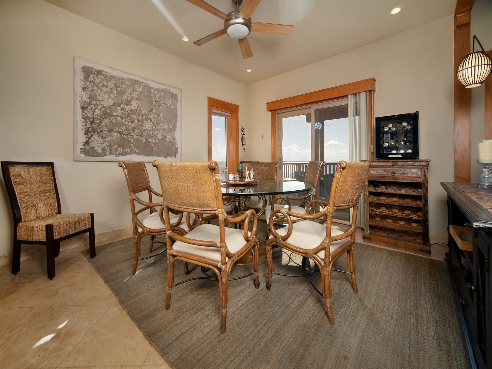 A dining area with a round table and six wicker chairs, a ceiling fan, and a large abstract painting on the wall.