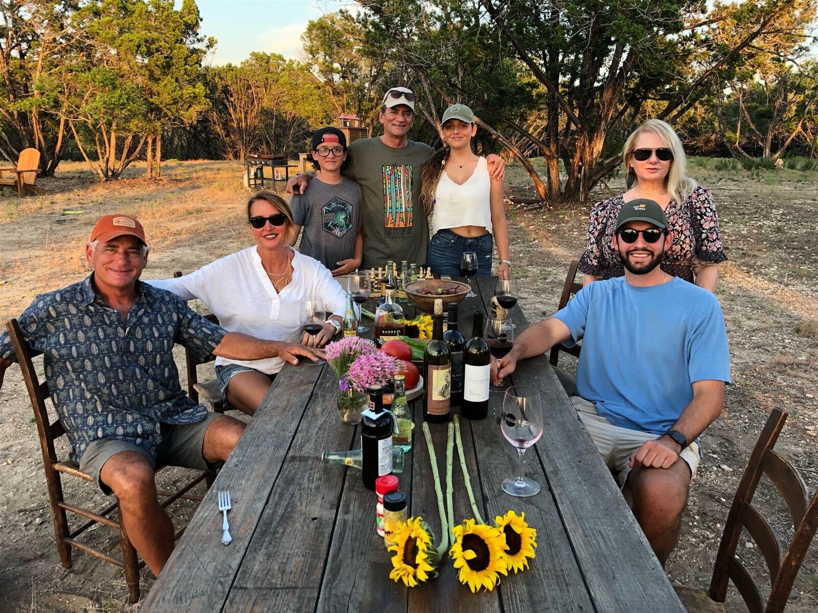 A group of people seated around a wooden table outdoors with various bottles and a bowl of food.