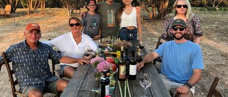 A group of people seated around a wooden table outdoors with various bottles and a bowl of food.