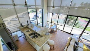A modern kitchen with a central island, a dining area with a table and chairs, and a large window with a view of parked cars and greenery outside. - Sunchase Resort Condo 20 ft glass view 4-min to water (South Padre Island)
