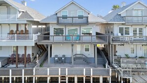 A multi-story waterfront house with a covered deck, outdoor seating, and a staircase leading to the upper levels.